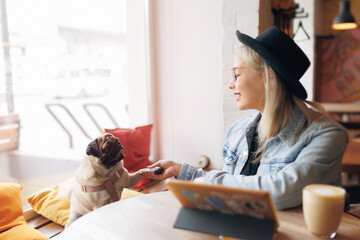 Concept friendship dog and people. Happy hipster student woman use tablet for study with pug. Freelancer work and drinking coffee in cafe