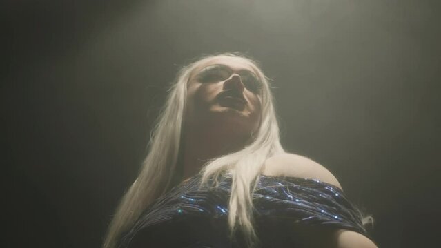 Low angle tilt up of fabulous drag queen wearing blond wig and emerald mini dress dancing seductively at camera in dark