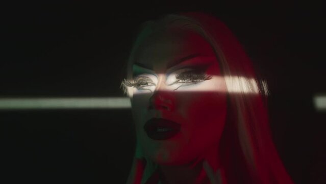 Closeup portrait of dramatic drag queen posing for camera in dark with horizontal light beam on her eyes with false lashes and wide black and blue arrows