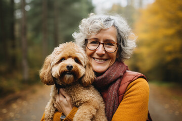 Portrait of mature woman holding her poodle dog in hands while posing for a shot in park - obrazy, fototapety, plakaty