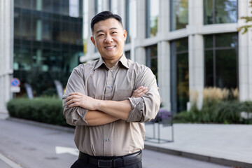 Portrait of a young Asian man standing near an office center with his arms crossed on his shoulders and looking at the camera with a smile