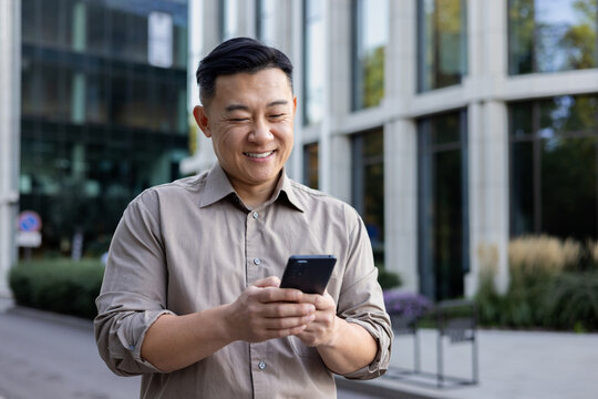 A smiling young Asian man holds a mobile phone in his hands and types messages, texts, calls. Standing smiling on the city street