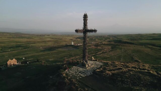 The largest iron cross in Armenia in the rays of the setting sun. On the background of high mountain Aragats in clear weather. Drone video. General plan, the camera moves in an arc.