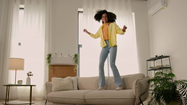 Active funny African American happy woman energetic girl teenager jump on sofa soft couch having fun fooling dancing vacation weekend jumping at home party rejoice moving day relocation dance music