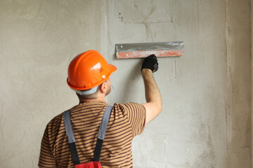 Rear view of handyman worker in overalls and protective helmet plastering concrete wall with putty...