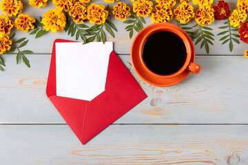 Obraz na płótnie Canvas Burgundy envelope with a white blank sheet of paper for text, an orange cup of black coffee and orange marigold flowers on a blue wooden table. Festive office desktop concept. Morning coffee cup.