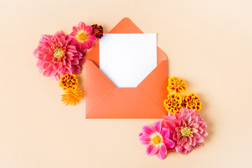 Orange envelope with clean white sheet for your text and autumn flowers pink dahlias and marigolds on a beige background. Nature trendy decorative design. Flat lay.