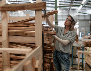 Confident female worker working in lumber warehouse of wooden furniture factory checking stock. Skilled carpenter inspector wears safety hardhat examining hardwood material for production facility.