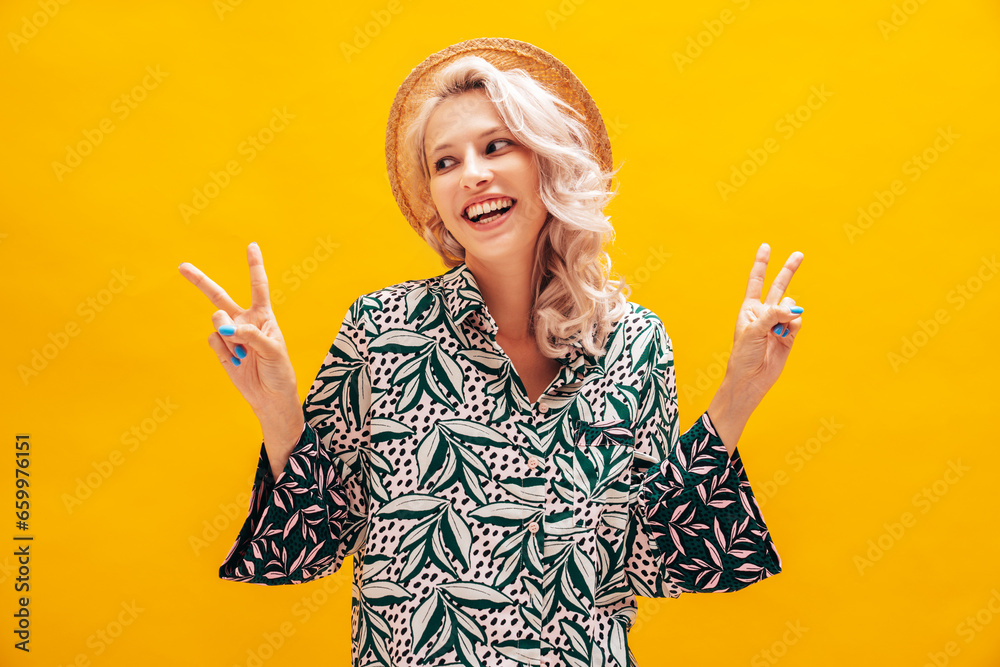 Poster Young beautiful smiling female in trendy summer hippie costume clothes. Carefree woman posing near yellow wall in studio. Positive blond model. Cheerful and happy. In hat. Shows peace sign