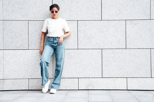 Young beautiful smiling hipster woman in trendy summer white t-shirt and jeans clothes. Carefree woman, posing in the street at sunny day. Positive model outdoors near wall. In sunglasses