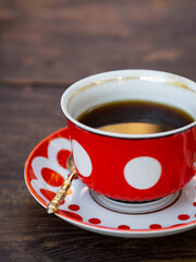 Black coffee in a red cup with polka dots and a saucer with a golden teaspoon on a wooden table.