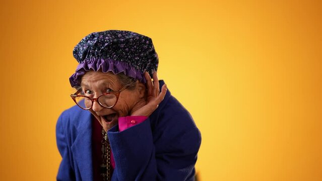 Funny portrait of smiling happy crazy toothless grandmother with wrinkled skin puts hand to ear to listen to a secret isolated on yellow background studio