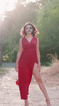 Vertical video, Beautiful sexy woman in a red evening dress and awesome Halloween makeup stands against the backdrop abandoned building. Greasepaint for Holiday on October 31. Slow motion.