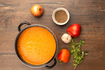 Top view of tomato puree soup in a pan and vegetables on a wooden background.