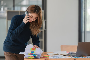 A businesswoman stands talking on her mobile phone while searching through documents on her desk at...
