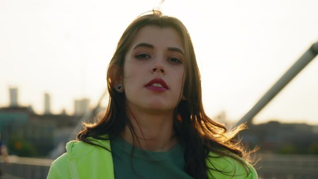 Portrait relaxed teenager girl standing outdoors in city looking into camera. Caucasian young female wearing light green hoodie backlit with warm sunset light, sunny summer evening, bright sun behind