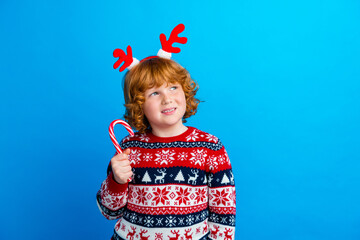 Photo of sweet dreamy little boy dressed ugly pullover horns licking x-mas caramel empty space isolated blue color background