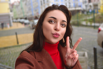 Charming lady in a brown autumn coat takes a selfie on the street with a peace sign