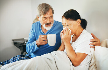 Sick senior woman in bed with husband blowing her nose with tissue for flu, allergies or cold. Love, comfort and elderly man in retirement hugging his ill wife in bedroom of modern home on a weekend.
