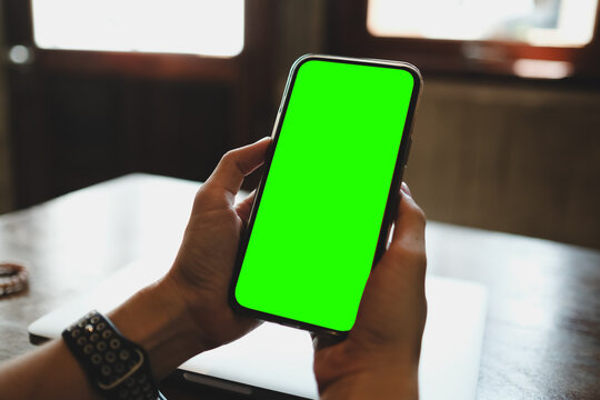 Mockup image of a business people holding smart mobile phone with blank green screen on vintage wooden table in modern cafe restaurant during meeting or lunch..