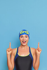 Happy girl in swimming suit and cap stands on blue background pointing her fingers up, professional sport concept, copy space