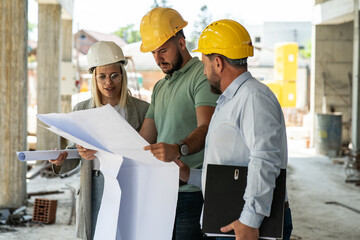 A female architect and construction managers work together on-site, discussing blueprints and...