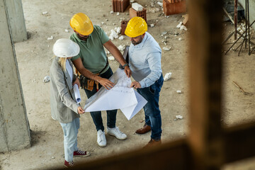 A female architect and construction manager work together on-site, discussing blueprints and...