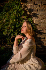 barefoot beautiful blonde woman in a white dress and wreath from green leaves sitting in ruins near...