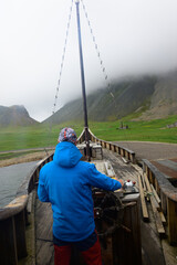 A captain of a boat is driving a boat. In the background are the misty mountains of Iceland.
