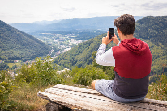 Hiking man sitting on a bench at a viewpoint enjoying taking photos of beautiful mountains and valley landscape with a smartphone