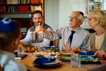 Jewish extended family lighting menorah during Hanukkah meal at dining table.