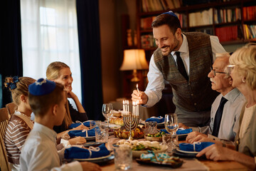 Happy Jewish man lighting candles in menorah while having meal with his family on Hanukkah.