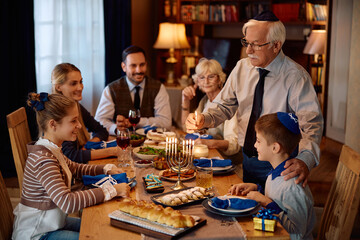 Jewish senior man lighting menorah during family meal at dining table on Hanukkah.