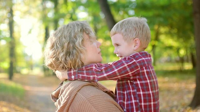 Close-up video portrait of a mother and son during a walk in the autumn park. Woman holds her toddler son in her arms and hugs him tenderly. A little boy is having fun and fooling around with his mom.