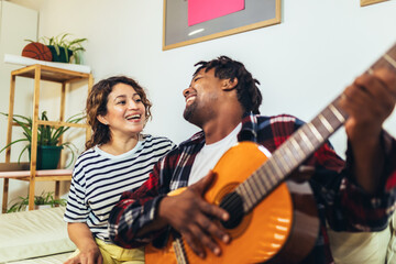 Happy young couple singing a song playing guitar on sofa in living room at home - Powered by Adobe