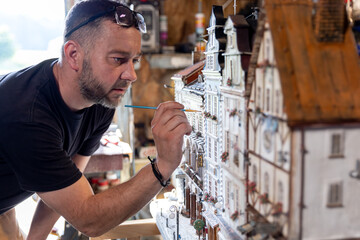 Modeller man working at scale model of miniature building in his workshop full of tools