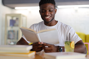 A happy young student in a university library, sitting with a book and a notebook, exudes...