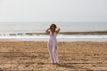 Young beautiful blonde woman in white dress is walking on the sand on the shore of the beach on a sunny day. The woman makes different body expressions. In the background the blue sea.
