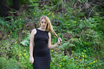 Young, beautiful blonde woman dressed in black walks through the forest in different postures and expressions. In the background ferns and yellow flowers. Concept expressions in nature.