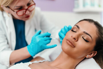 Beautician is contouring the woman's cheekbones with hyaluronic acid filler. Hyaluronic acid filler is injected by needle or cannula. Face contouring concept. - obrazy, fototapety, plakaty