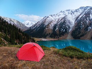 Camping with a red tent in the highlands on the shore of a beautiful lake. Panoramic views of snow-capped peaks and turquoise lake