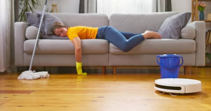 Tired woman lies on sofa while a robot vacuum cleaner does the cleaning