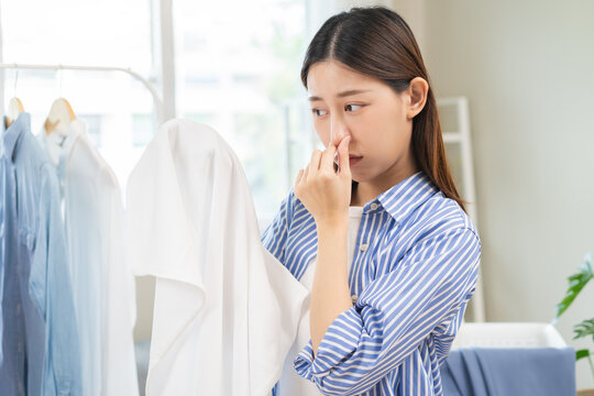 Asian young housewife woman having bad smelling clothes hand holding breath nose with finger, sniff smelly dirty stinky musty, look disgusting from clothes after washed, laundry out of machine at home