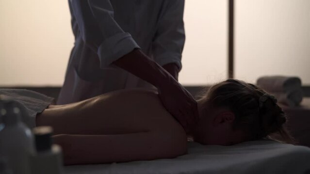 Medium shot. Masseur, massage specialist giving neck massage to his patient. Silhouettes of a woman and a man in the massaging room, spa procedure.