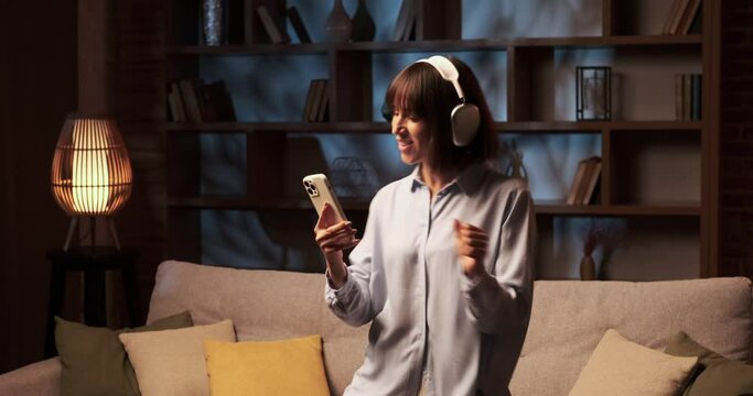 Woman wearing headphones and with phone in hands moves gracefully, lost in the rhythm of the music playing on phone. Her body sways to the beats, and face radiates pure joy.