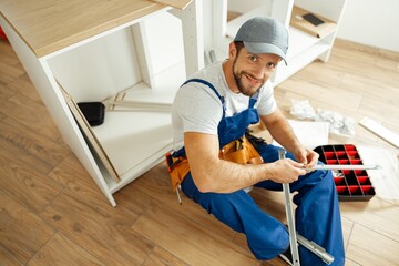High angle view of handyman in uniform smiling at camera while sitting on the floor and assembling drawer in kitchen