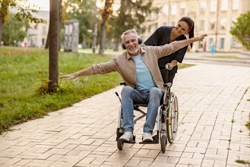 Joyful mature disabled man in wheelchair wearing headphones having fun during a walk in the city assisted by lovely young nurse