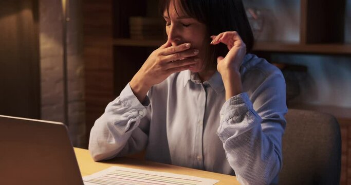 Weary woman in late hours of the evening stifles a yawn as she continues to work diligently. The fatigue is evident in eyes, yet dedication keeps going.