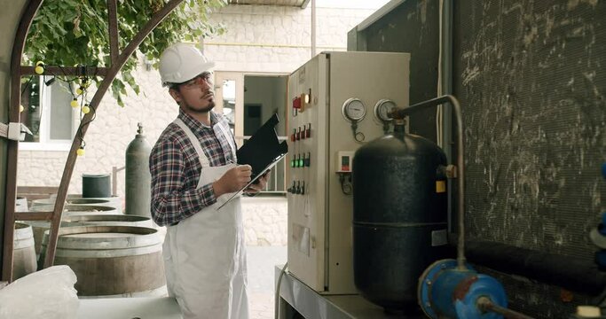 Male wine maker checking quality of beverage distillation process indoors and write down the dates in the diary. Alcohol production concept.Controls the quality of production equipment in wine factory