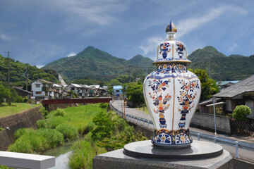 A porcelain vase in the traditional Japanese Imari style - red, blue and gold colours, picturesque landscape of the town of Arita in the background. - obrazy, fototapety, plakaty